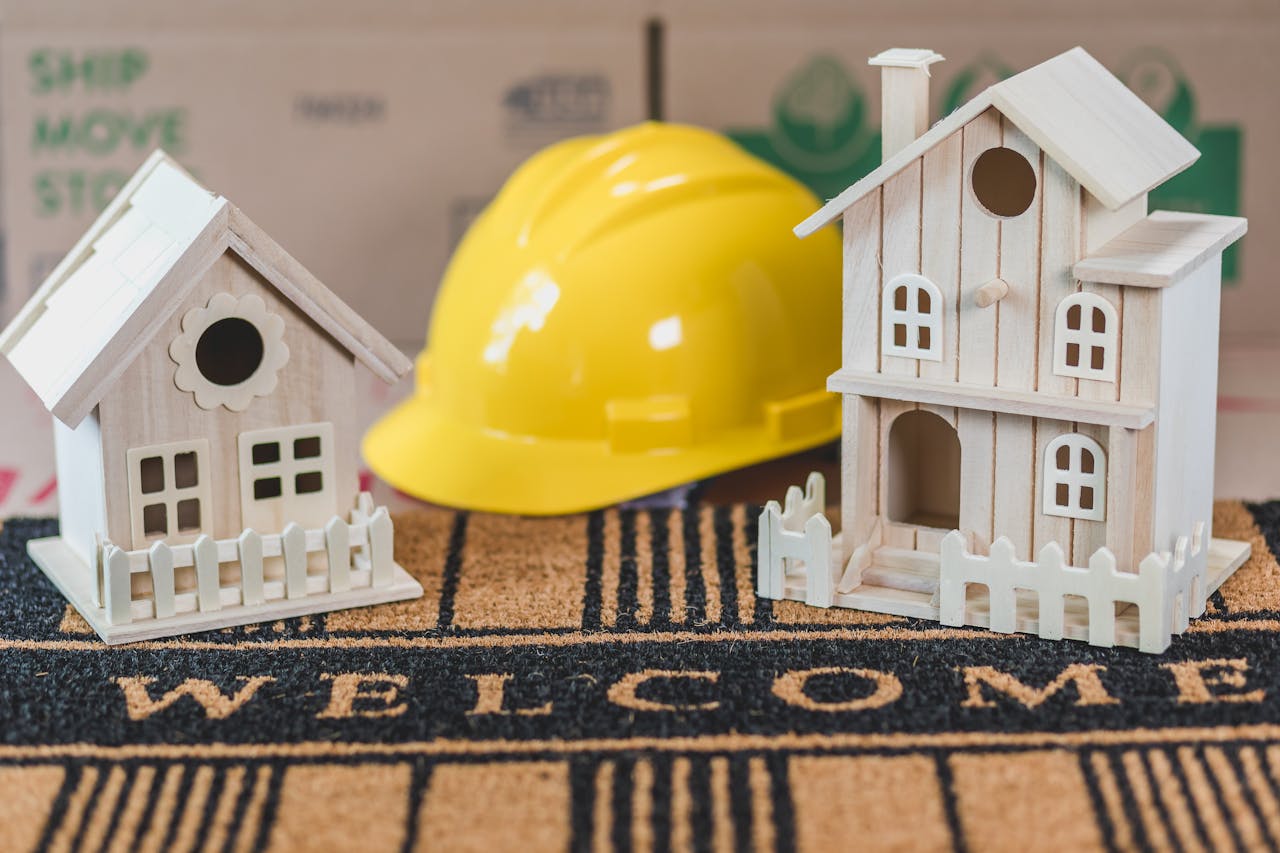 Miniature wooden houses on a welcome mat with a yellow hard hat in the background, symbolic of home building or construction.